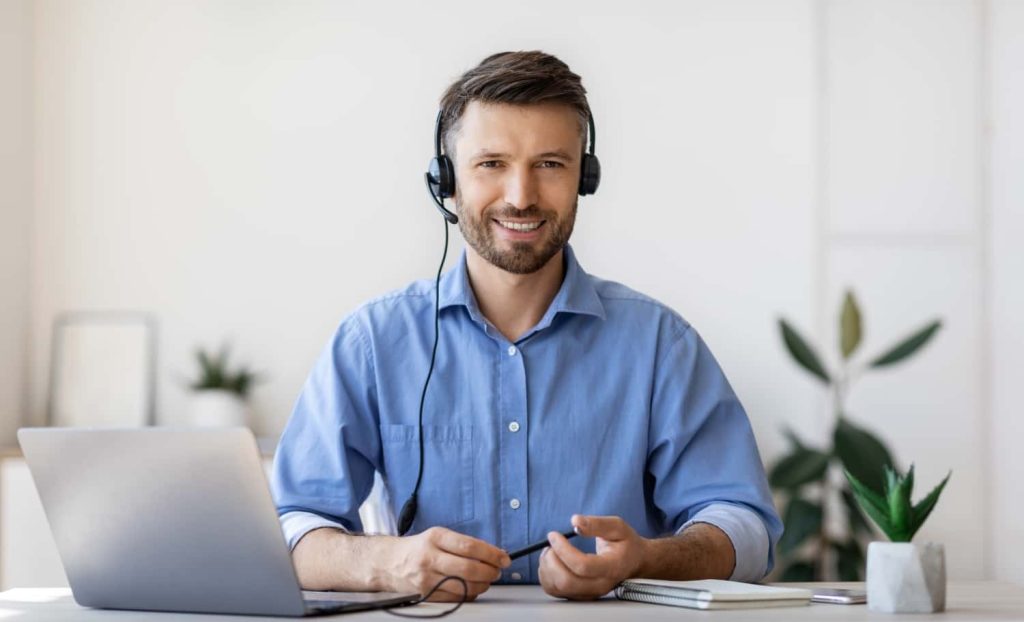 A smiling man wearing a headset sits at a desk with a laptop, ready to assist clients with tax and accounting services. The background features plants and a bright, modern workspace.