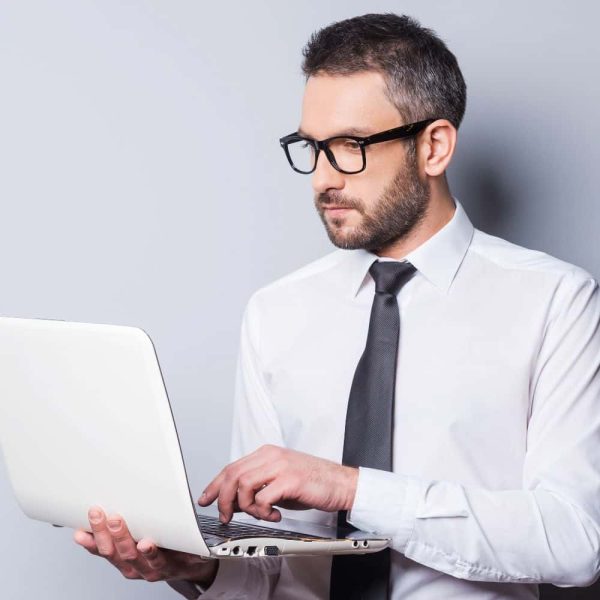 A man in a white shirt and tie is working on a laptop, wearing glasses and looking focused. The background is a light gray color.